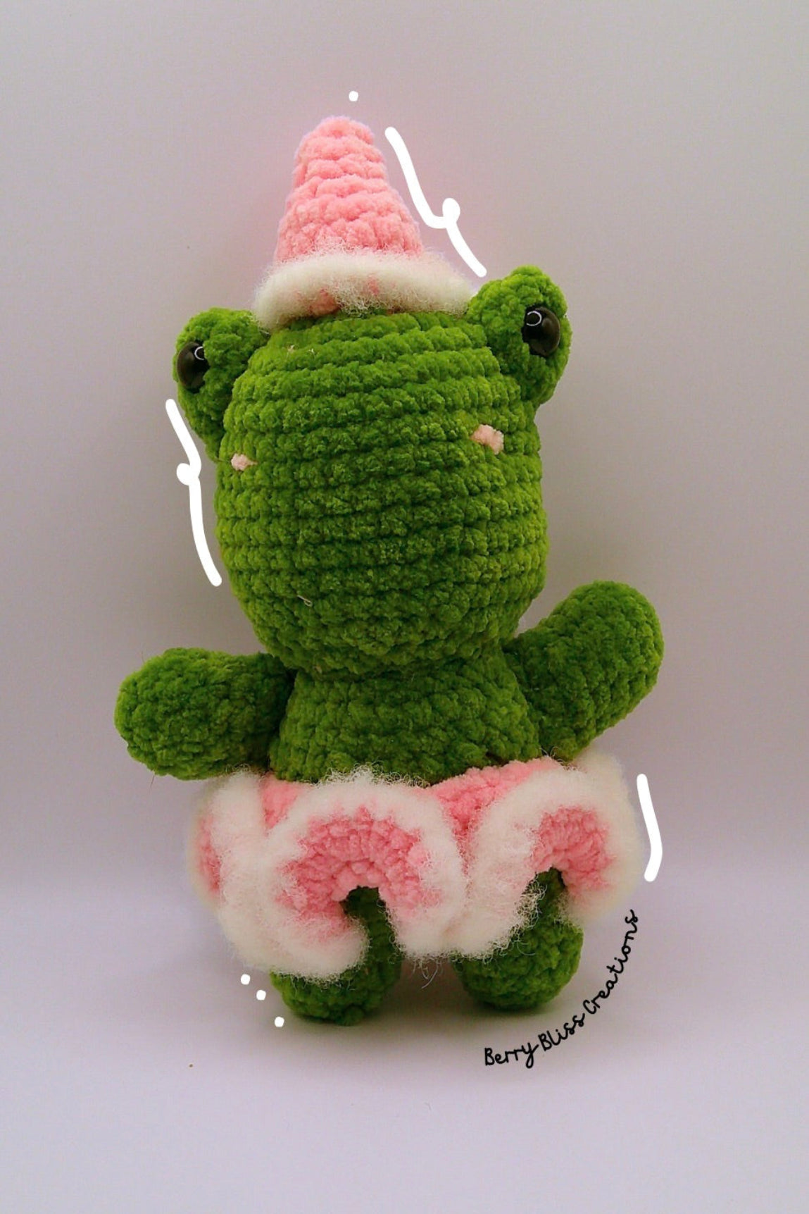A green crocheted plush frog toy wearing a pink party hat with white trim and a pink-and-white skirt.