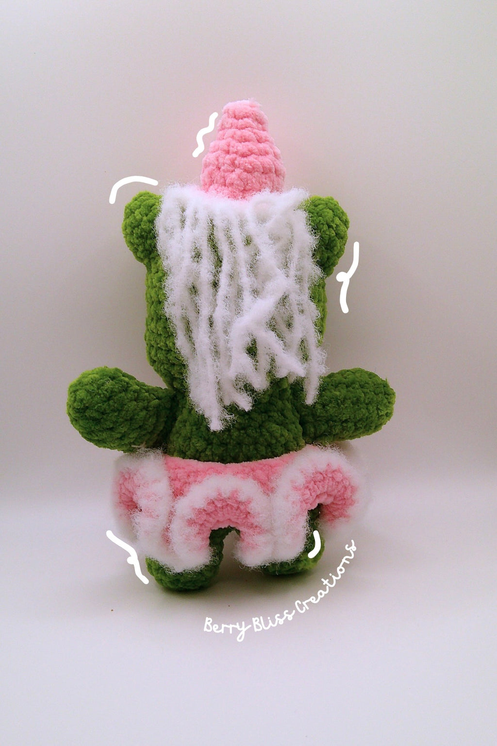A handmade crocheted plush toy shaped like a green character wearing a pink hat and a pink-and-white ruffled skirt, with white yarn hair.