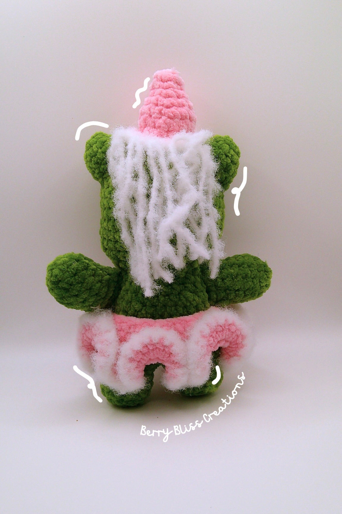 A handmade crocheted plush toy shaped like a green character wearing a pink hat and a pink-and-white ruffled skirt, with white yarn hair.