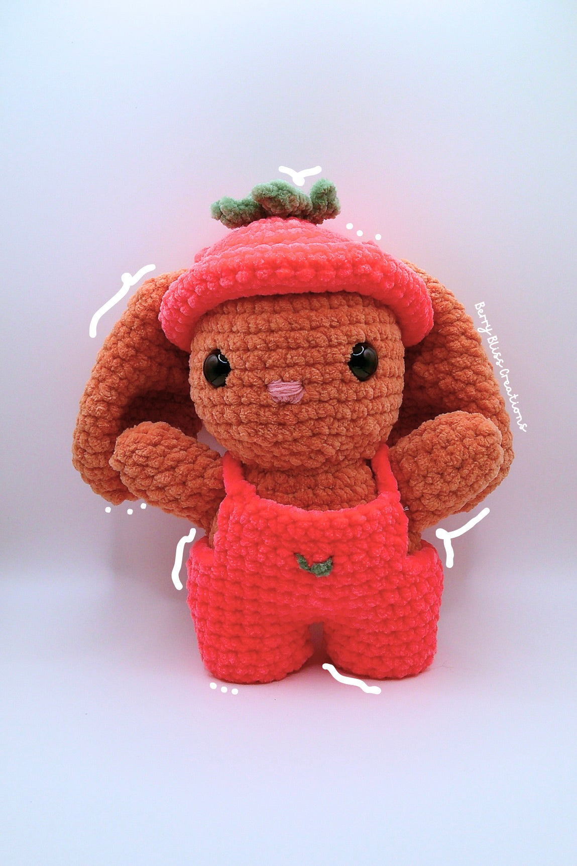 A crocheted plush bunny doll with a strawberry theme, featuring a red hat with green leaves, a pink nose, and red overalls with a small green leaf detail.